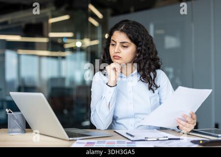 Young professional woman concentrating, carefully analyzing documents and reviewing information while sitting at her desk in a modern corporate office environment Stock Photo
