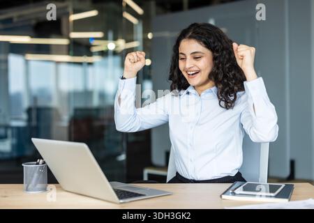 Excited young businesswoman clenching fists in triumph, celebrating business victory or achievement while looking at her laptop screen in a modern office workplace Stock Photo