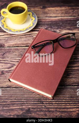 Vertical still life of eyeglasses on a closed hardcover book with a cup of coffee on rustic wood. Concept of reading, study and work from home. Stock Photo