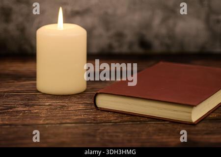 Lit candle next to a closed red book on a rustic wooden table. Minimal cozy background with copy space for text and branding. Stock Photo