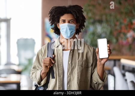 Safety Measures. Black Guy Wearing Medical Mask Showing Blank Smartphone At Cafe Stock Photo