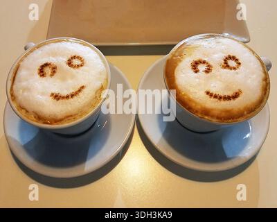 Two Cappuccino Coffee cups with froth sprinkled cocoa and cinnamon in the shape of smiley faces. Stock Photo