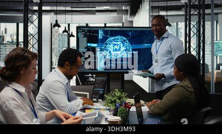 Programmer gives presentation to executives, implementing AI chatbot reducing work volume. Startup office IT expert analyzing business insights for management using artificial intelligence, camera A Stock Photo
