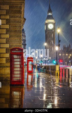 London by night with rain and storm in front of the Big Ben Tower with red telephone booths and water puddle reflections on the street Stock Photo