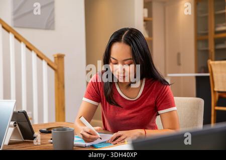 Asian woman sitting at home table writing on blue sticky notes with pen wearing red T-shirt Stock Photo