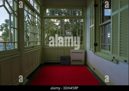 Kobe, Japan - October 30, 2023: A sunroom in the Moegi House features large geometric windows, green wooden shutters, a red carpet, a wall-mounted heater, and a metal box, with trees visible outside. Stock Photo