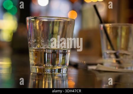 A close-up view of a clear glass half-filled with liquid sitting on a reflective dark table surface at night. The background features colorful soft bo Stock Photo