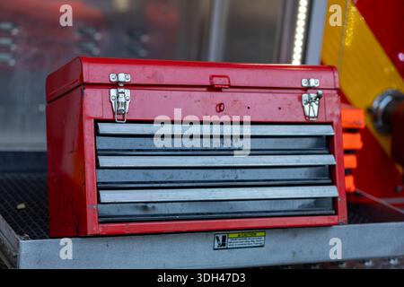 A close-up view of a worn red metal industrial toolbox chest with multiple drawers and silver latches resting on a textured silver diamond plate metal Stock Photo