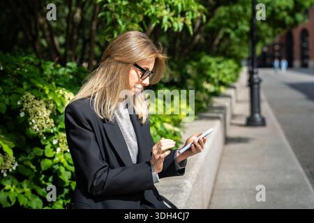 Fashionable businesswoman sitting outdoors in sunny weather and enjoying scrolling on smartphone. Stock Photo
