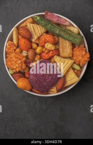 A bowl of assorted snacks including nuts, raisins, and potato chips. The bowl is on a dark surface Stock Photo