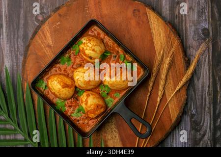 A black pan with eggs masala / egg curry on a wooden table. The eggs are arranged in a row and the sauce is spread out over the pan. The scene has a w Stock Photo
