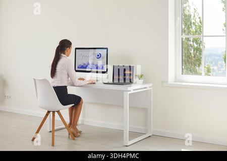 Businesswoman analyzing charts at home office with desktop monitor and laptop Stock Photo