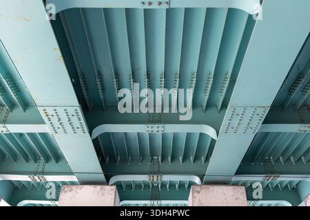 Perspective view of the underside of a teal-painted steel bridge, featuring riveted girders, repeating ribs, and structural spans. Ideal for architect Stock Photo