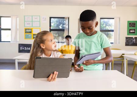 Classmates schoolchildren holding tablets in classroom, boy in green T-shirt talking to seated girl Stock Photo
