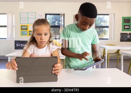 Diverse students sitting at white table in bright classroom using tablets in folio cases Stock Photo
