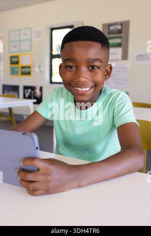African school-age boy wearing mint green T-shirt holding gray tablet at white desk in classroom Stock Photo