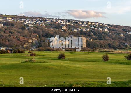 Harlech, Wales, March 06, 2024: View across a green golf course toward the hillside town, with terraced houses and buildings catching warm evening lig Stock Photo