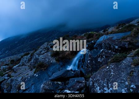 Waterfall on the Afon Lloer flowing over rocks on the slopes of Pen y Ole Wen under low cloud Stock Photo