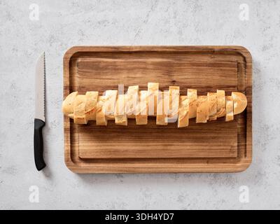 Baguette bread loaf partially sliced on a wooden cutting board with a serrated knife, seen from a top view Stock Photo