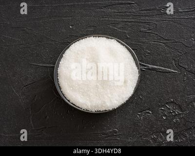 White crystal granulated sugar in a small glass bowl on dark textured background, overhead flatlay studio shot, close-up Stock Photo