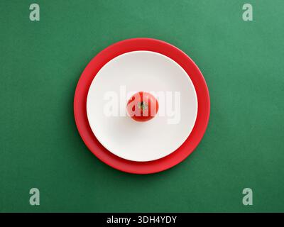 Tomato resting on white and red plates on a green background. Minimalist top view composition Stock Photo