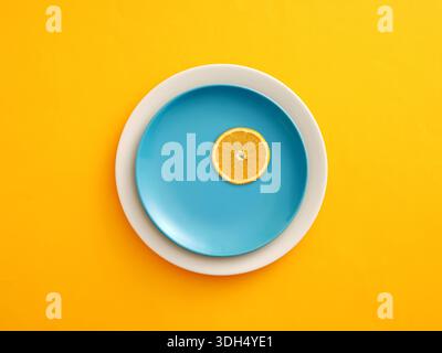 Orange slice resting on a blue ceramic plate, stacking on a white plate. Bright yellow background creating a minimalist setting Stock Photo