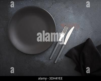 Empty black plate and cutlery on a dark textured background, creating a minimalist table setting for dinner or a meal Stock Photo