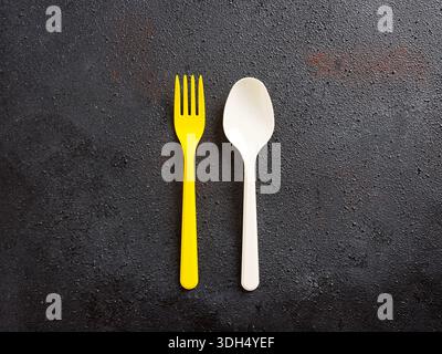Yellow plastic fork and white plastic spoon creating a contrast on a dark background, representing disposable cutlery Stock Photo