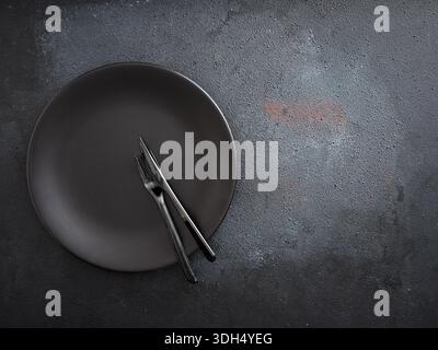 Empty black plate with matching fork and knife resting on a dark, textured background, creating a minimalist food concept Stock Photo