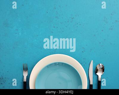 Empty dinner setting with blue plate on a beige plate, fork, knife, and spoon arranged on a vibrant blue background Stock Photo