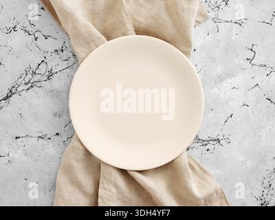 Empty round plate with a beige linen napkin on a gray marble table, creating a minimalist flat lay setting Stock Photo
