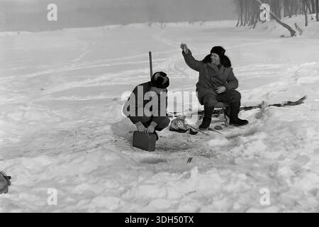 Authentic archival photograph from the 1960s capturing a joyful moment of success during an ice fishing trip near Raigorodok, Donetsk region. Two men in warm winter coats and fur hats are pictured on the frozen river. The man on the right, sitting on a box, triumphantly holds up a small fish he has just caught, smiling broadly. His companion crouches nearby, attending to a tackle box. Fishing gear lies on the snow nearby. This atmospheric black and white image documents the camaraderie and simple pleasures of peaceful life in the Donbas region before the war. Stock Photo