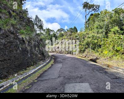 A wide-angle view of a paved road curving sharply past a dark, towering rock face. The area is surrounded by dense tropical foliage and tall trees und Stock Photo