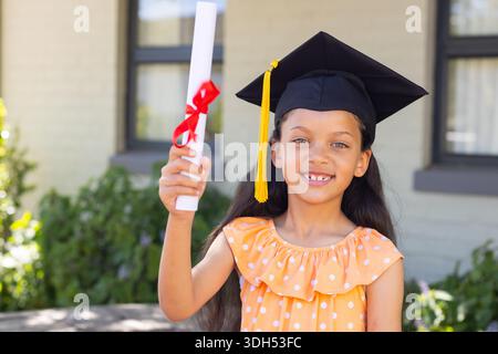 African American child wearing mortarboard and orange polkadot dress holding diploma in front yard Stock Photo