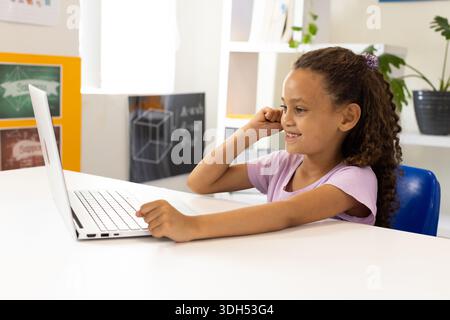 African American girl sitting at table using silver laptop, looking toward screen, hand near ear Stock Photo