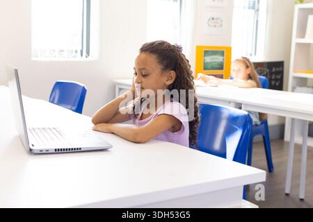 Diverse female students working on laptop at school classroom with blue chairs and bulletin board Stock Photo