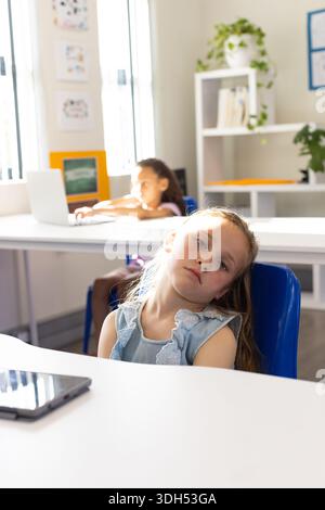 Diverse girl classmates sitting in classroom child leaning on blue chair tablet peer using laptop Stock Photo
