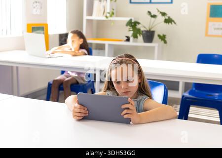 Diverse female children sitting in classroom, front child holding tablet, back child using laptop Stock Photo