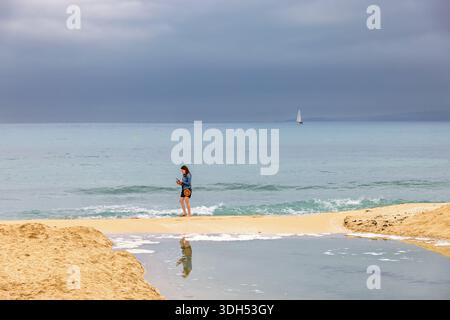Alone woman walking by the sea on a sand beach and watching the mobile phone Stock Photo