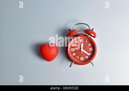 Red heart and black alarm clock on blue symbolic pairing suggesting love’s connection to time, urgency, or enduring emotional presence. Stock Photo