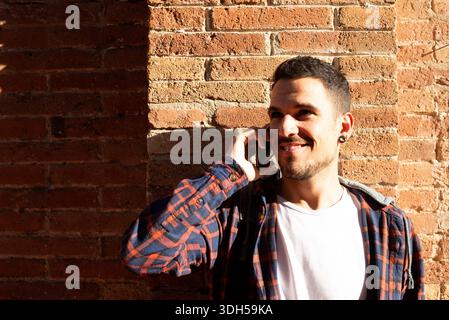 Adult caucasian man leaning against a brick wall while talking on a smartphone. He smiles, wears casual clothes, and stands outdoors in sunlight. Stock Photo
