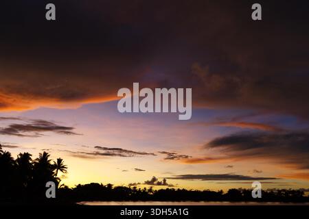 Dramatic sunset over Viti Levu, Fiji, with silhouetted palm trees beneath layered storm clouds glowing at dusk. Stock Photo