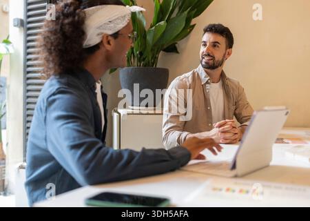 Two professionals engaging in a collaborative discussion at a coworking space. One is using a tablet while both are surrounded by a modern, creative e Stock Photo