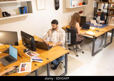A diverse group of coworkers collaborating in a modern coworking space. Individuals are focused on tasks using laptops at adjacent desks, promoting te Stock Photo
