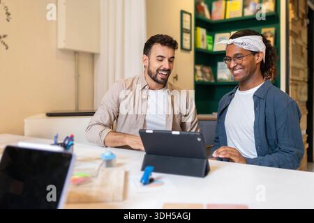 Two coworkers are happily collaborating at a coworking space. They are seated at a table, engaged in a discussion, with a tablet in front of them in a Stock Photo