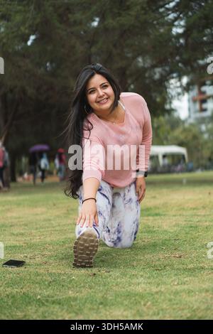 A cheerful woman stretches in a lush park, enjoying outdoor activities in a vibrant urban environment. Capturing the essence of healthy living in Lati Stock Photo