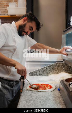 A skilled pizza maker diligently spreads tomato sauce on dough in a busy pizzeria. His focus and technique highlight the craftsmanship involved in mak Stock Photo