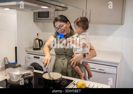 A mother balances cooking and childcare, holding her son while stirring food on the stove. She chats on the phone, demonstrating multitasking in a mod Stock Photo