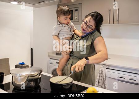 A mother skillfully balances her toddler on one arm while cooking and talking on the phone in a modern kitchen, showcasing multitasking and parenting Stock Photo