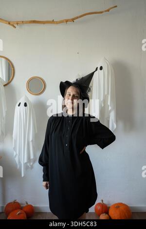 A cheerful woman dressed as a witch poses amidst whimsical ghost decorations and pumpkins, capturing the festive Halloween spirit in a creatively deco Stock Photo